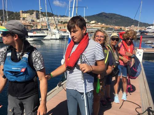 Un groupe de pratiquants déficients visuels marchent sur un ponton et s'apprêtent à embarquer à bord d'un voilier